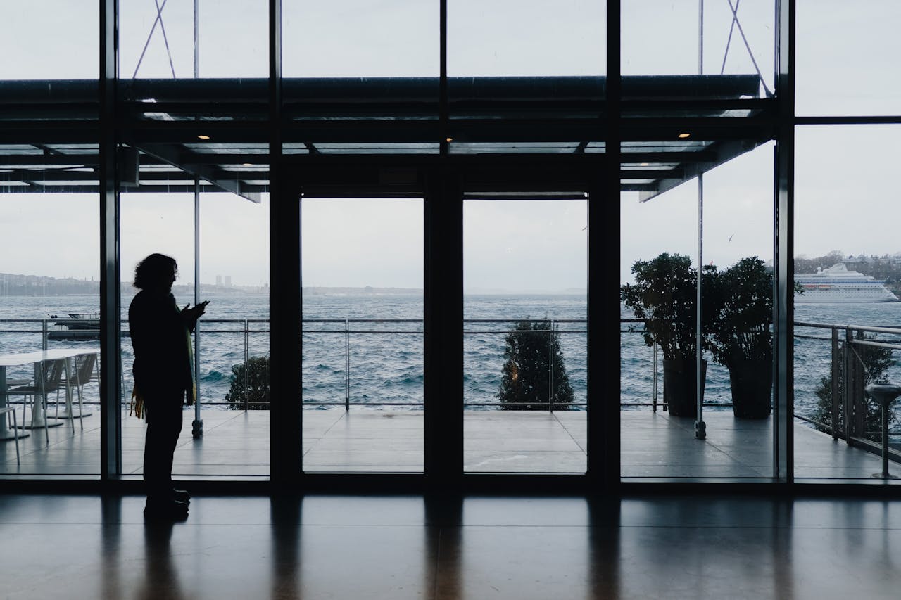 Home Silhouette of a person indoors by a waterfront, viewed through glass panels on an overcast day.